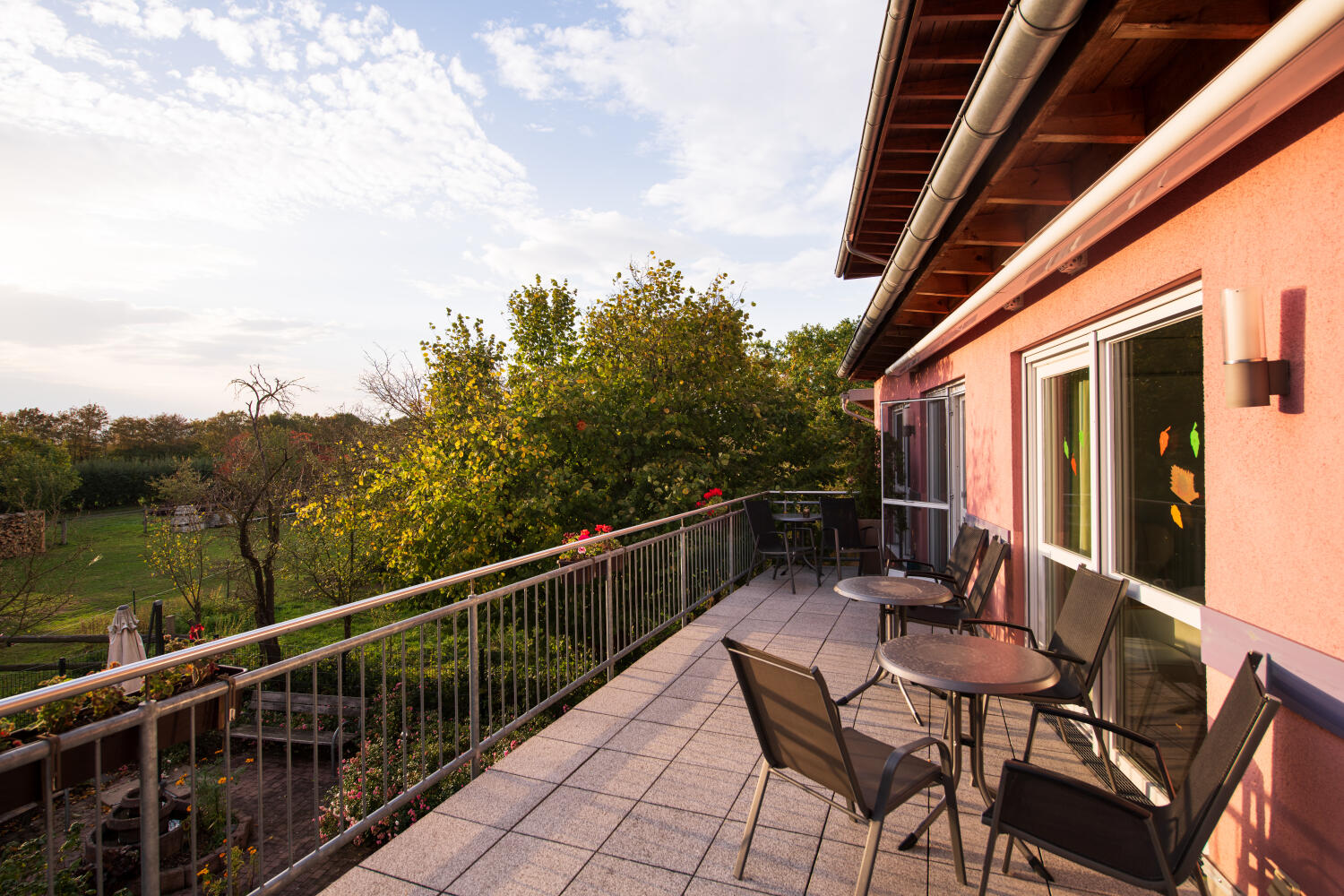 Balkon mit runden Tischen und leichten Stühlen. Links übers Geländer Blick in den grünen Garten.