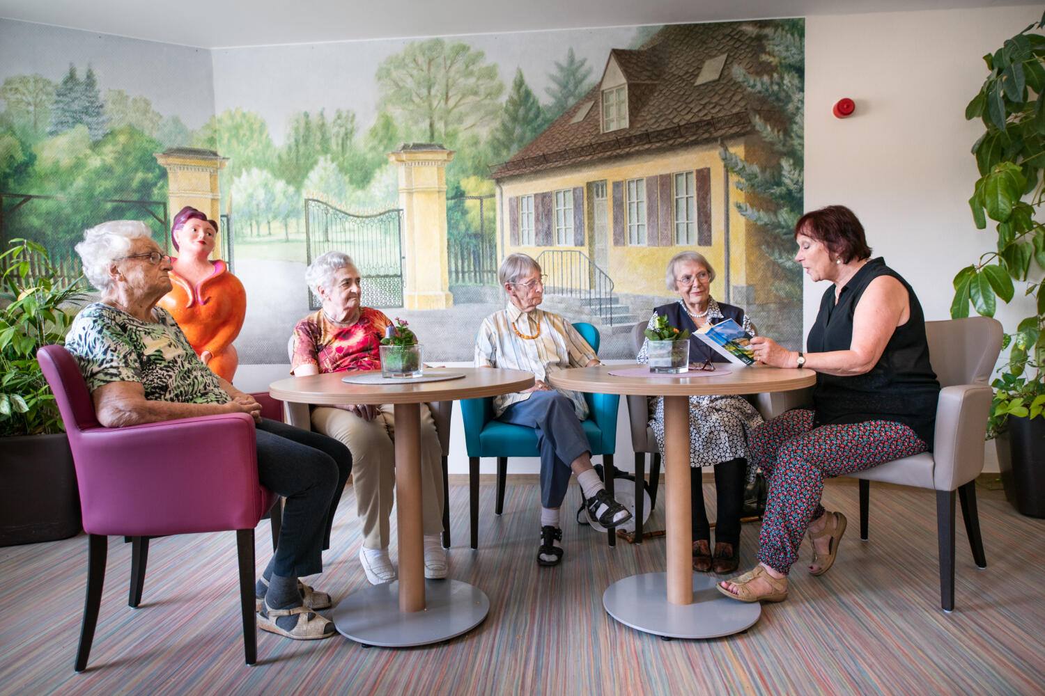 In einer Zimmerecke mit zwei runden Tischen sitzen 4 aufmerksame ältere Damen, die einer Vorleserin zuhören. Auf der hinteren Wand ist ein imposantes, buntes Wand-Bild. Es zeigt ein Häuschen zu einem Parkeingang mit schmiedeeisernem Tor. 