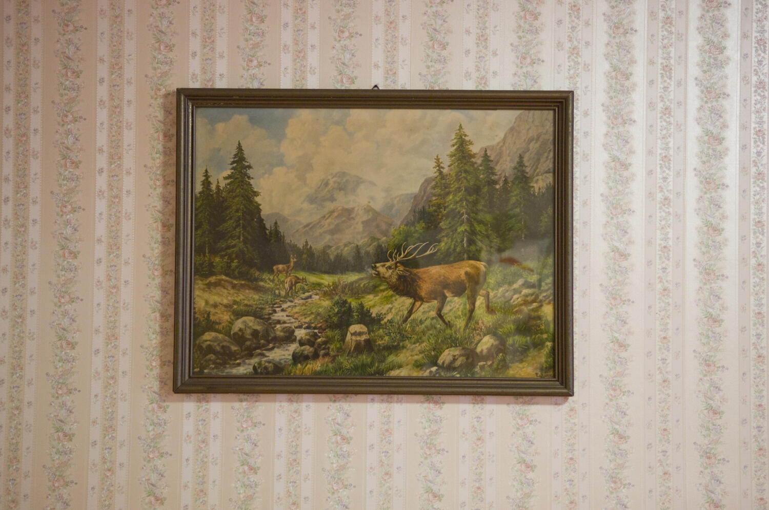 Auf einer zart geblümten Wand   in Streifen hängt ein großes, dunkel gerahmtes Bild einer Gebirgslandschaft, einem Bach und dem  "berühmten röhrenden Hirsch" samt Ricke und Kitz.