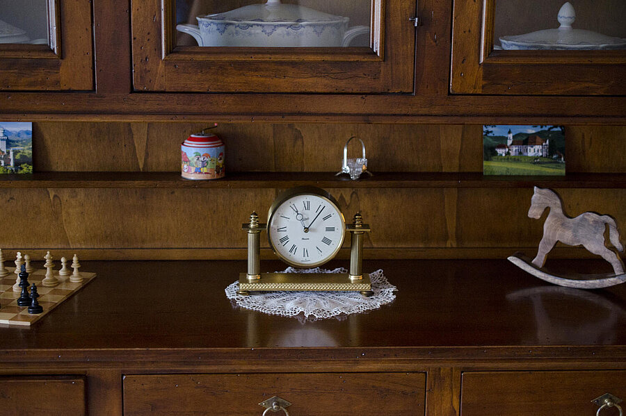 Auf dem Vorderteil einer Vitrine, in der man Geschirr erkennen kann, stehen neben einer Uhr ein Schachbrett, ein kleines Schaukelpfer, zwei Postkarten und eine bunte Dose.