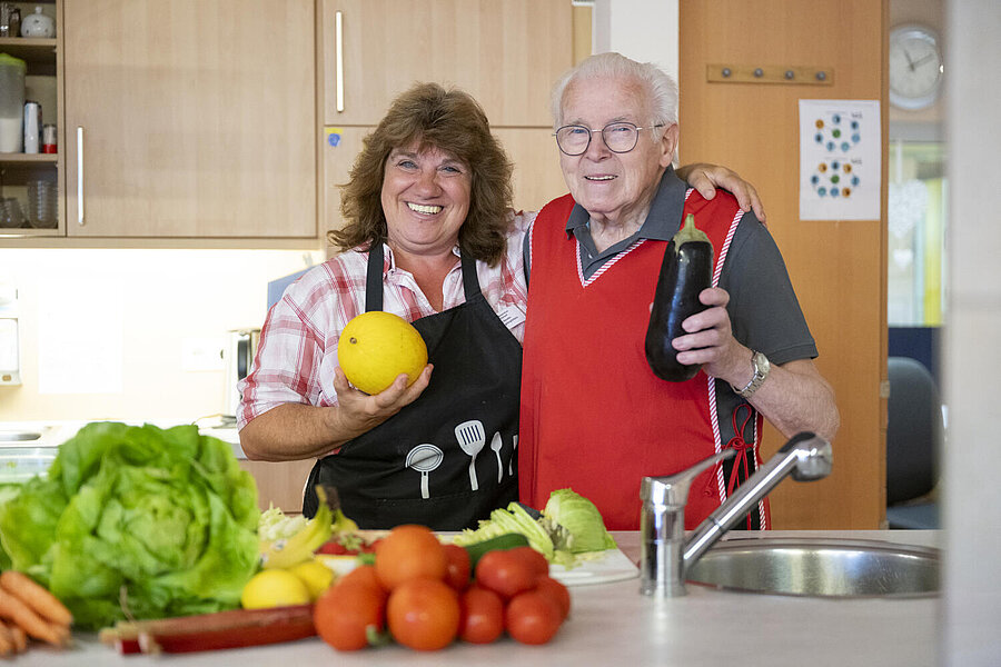 Älterer Herr und deutlich jüngere Frau zeigen lächelnd und stolz Obst und Gemüse.