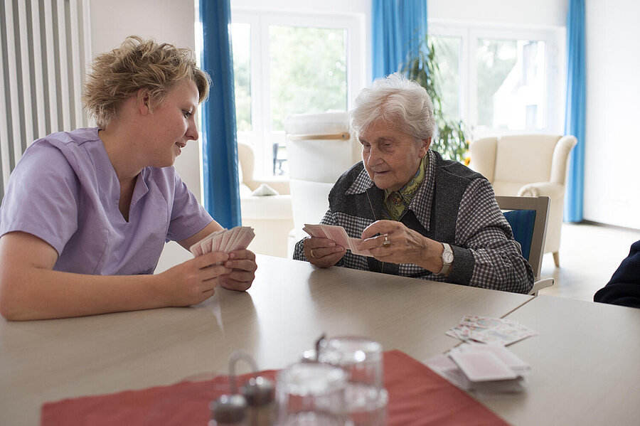 Eine ältere Bewohnerin und ein jüngere Betreuerin spielen an einem Tisch ein unbekanntes Kartenspiel. Vor Ihnen  stehen auf einer roten Serviette leere Gläser und ein Gewürzständer.