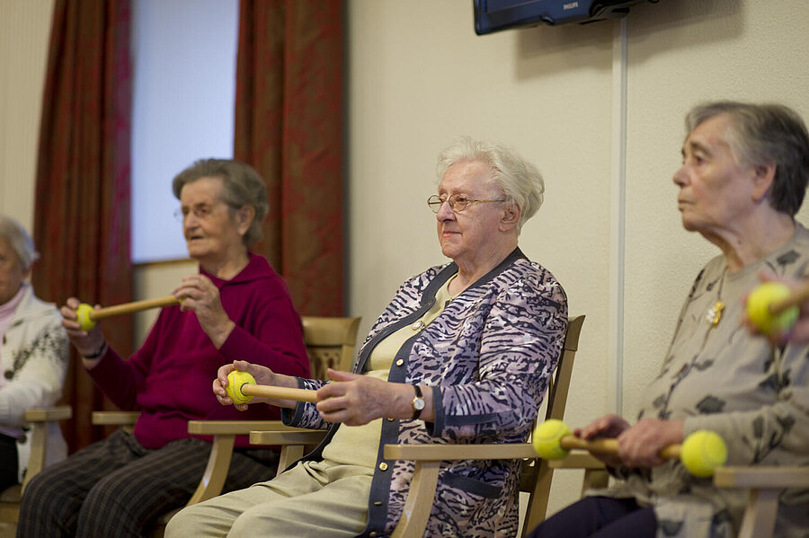 Drei ältere Damen sitzen auf Stühlen und halten Stöcke in den Händen, auf die links und rechts je ein Tennisball aufgesteckt ist. Sieht wie "Hockergymnastik" aus.