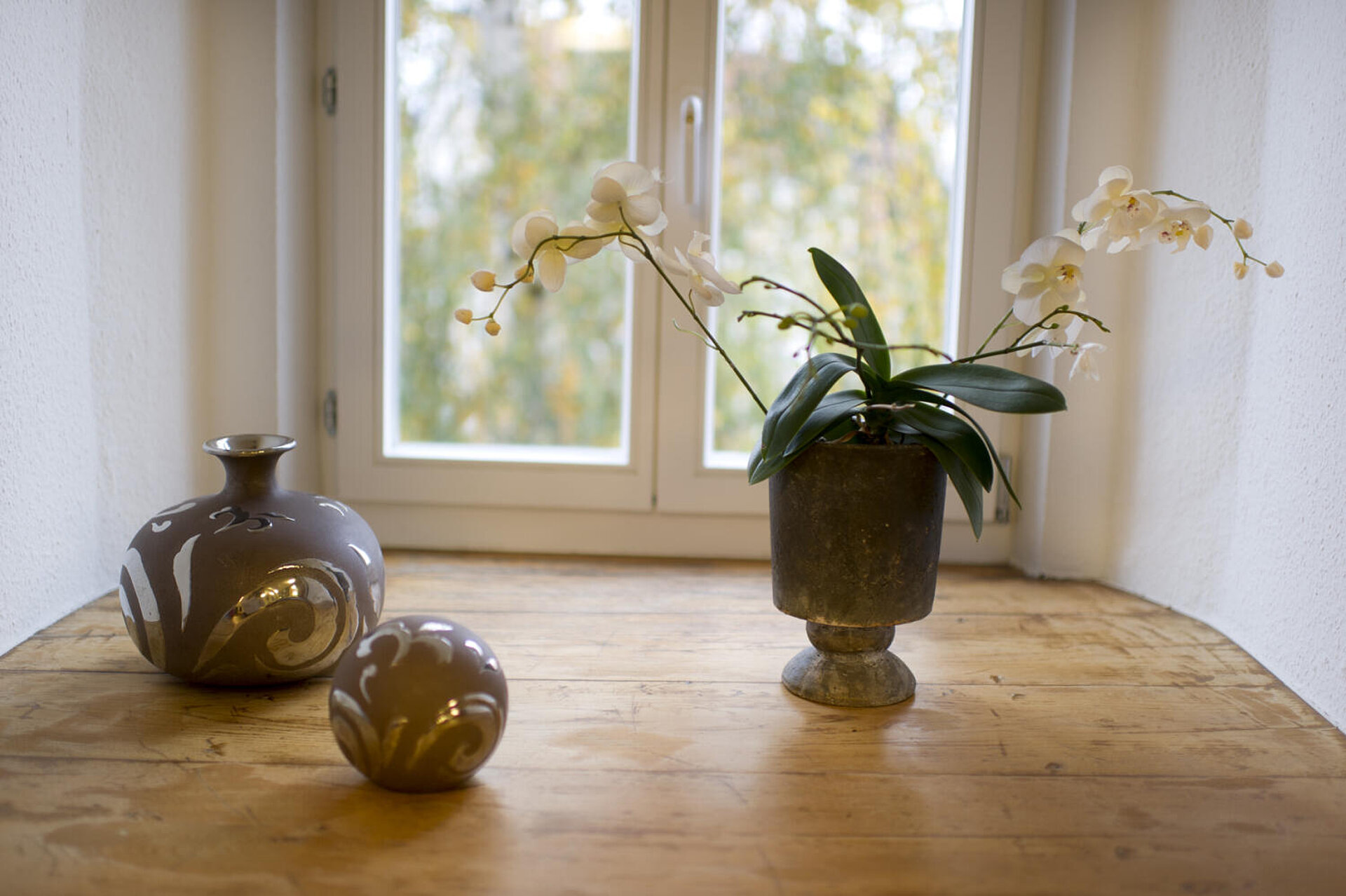 Auf dem hellbraunen Dielen-Fußboden vor einer Glastür stehen drei Keramikvasen:  Eine zylinderförmige mit Blumen, eine leere Bauchige, die Dritte ist ein Kugel.
