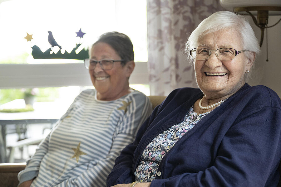Zwei nebeneinander vor einem Fenster sitzende alte Damen strahlen den Fotografen an. Ein ansteckendes Lachen.                   