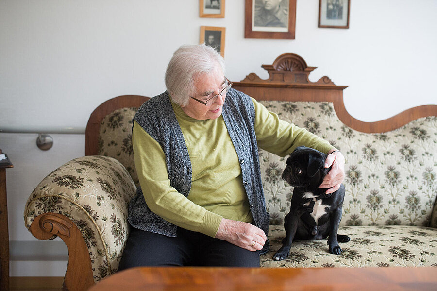 Auf einem zweisitzigen Kanapee mit buntem Bezug sitzt eine ältere Dame, die einen neben ihr sitzenden, kleinen, schwarzen Hund  am Kopf streichelt. Dem scheint es nicht ganz geheuer zu sein, wie sein Blick samt gespannter Körperhaltung verrät.