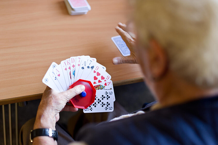 Vor einem Tisch sieht man von  rückwärts, etwas  verschwommen eine Kartenspielerin oder Kartenspieler. Mit der linken Hand mit Armbanduhr werden mit einem roten Kartenhalter 12 ausgefächerte Karten gehalten. Ein Stapel und eine einzelne Karte liegen auf dem Tisch.