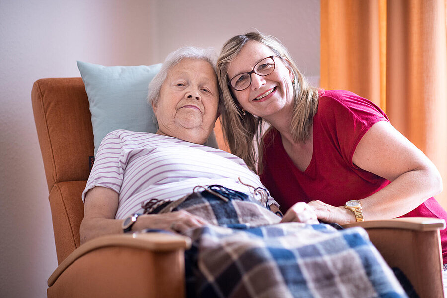 Eine ältere,  im Sessel ruhende Bewohnerin erhält gerade eine lächelnde Zuwendung von einer Betreuerin, die sich der Bwohnerin zuneigt.