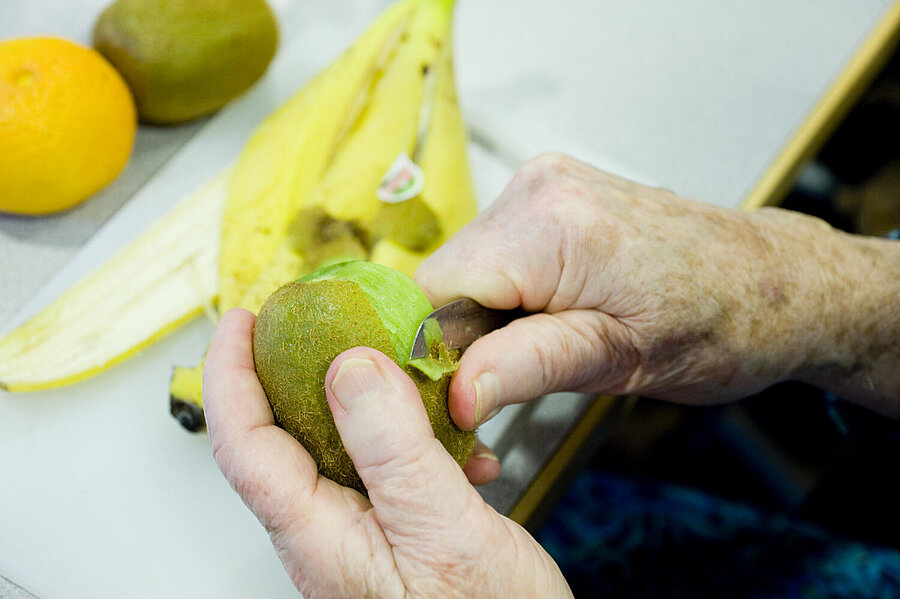 Auf einer Tischplatte liegen eine Banane, eine Kiwi und eine Pampelmuse oder sehr helle Orange. Zwei Hände einer nicht weiter erkennbaren Person schälen gerade eine Kiwi-Frucht.