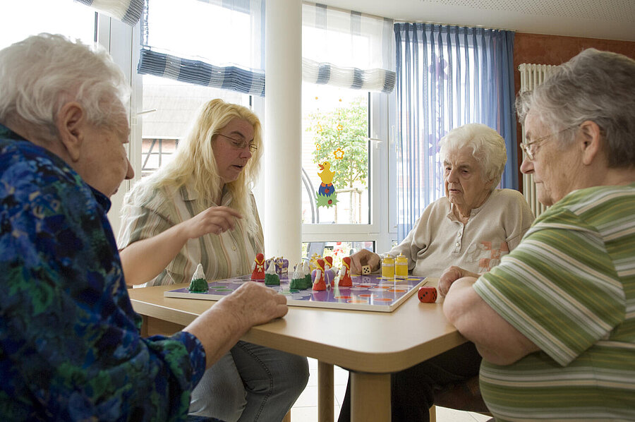 An einem kleinen Tisch sitzen vier Frauen, unterschiedlich gekleidet, die engagiert in ein Würfelspiel  vertieft sind, das mit vielen bunten, kleinen Figuren auf einem Tablett gespielt wird.