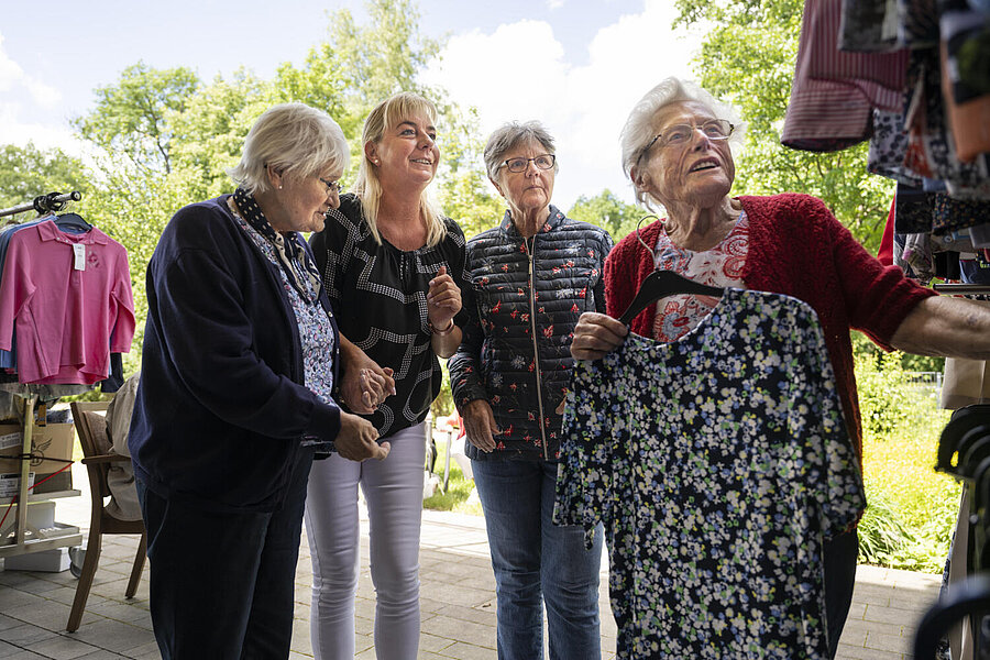 Drei ältere Damen, eine von einer jungen Betreuerin gestützt, betrachten Kleidungsstücke, die auf zwei fahrbahren Regalen hängen. Eine der Damen hält eine bunte Bluse auf einem Bügel in der Hand.
