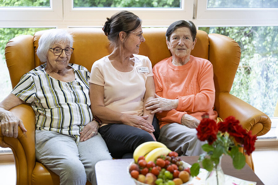 Auf beige-gelbem Sofa drei Damen, die sich offensichtlich gut verstehen. Vor ihnen auf einem Tisch rote Rosen und volle Obstschale.
