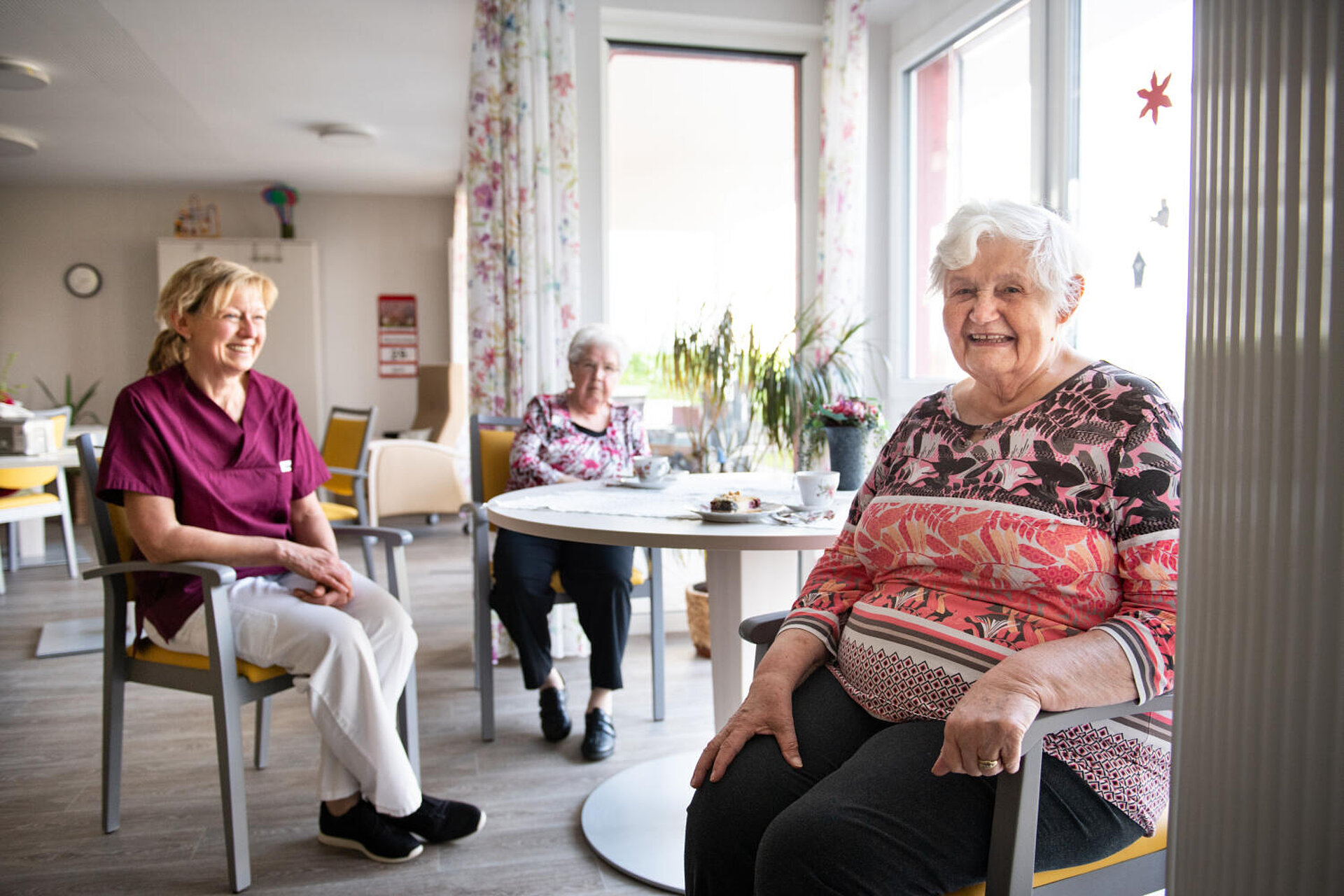 In der rechten Fensternische am runden Tisch mit Blumen sitzen zwei ältere Damen beim Kaffeetrinken. Ein jüngere Betreuerin sitzt - ein klein wenig abgesetzt - herzhaft lachend dabei.