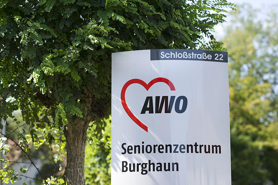 Das Bild wird eingenommen von dem bekannten, vereinheitlichten "AWO-Standschild" mit "AWO-Logo" und der Aufschrift "Seniorenzentrum Burghaun" und dem Zusatz am oberen Rand: Schloßstraße 22. Alles vor sattem Grün.