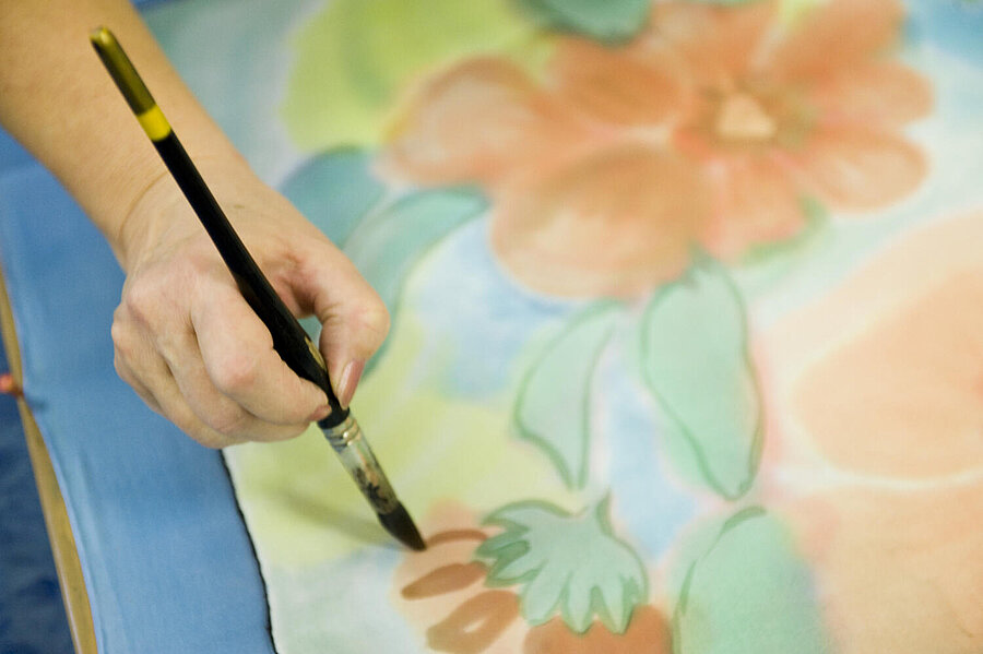 Eine Hand mit Pinsel - von oben gesehen - malt Blumen auf weißem Papier.  Das Blatt liegt auf einer blauen Unterlage.
