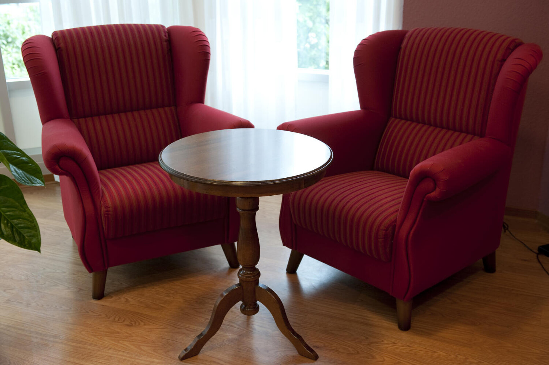 In einer Zimmerecke stehen um einen kleinen, runden, leeren Tisch zwei rote Plüschsessel.