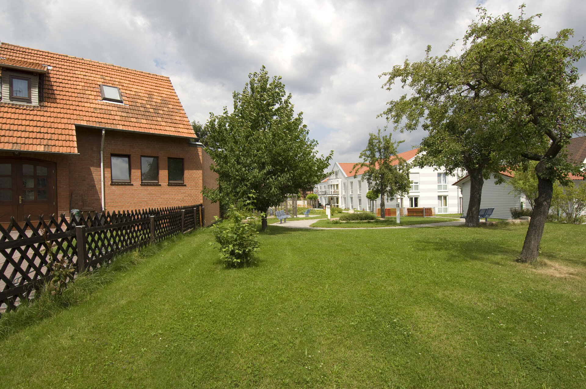 Vom Rande einer großen Rasenfläche mit mehreren großen Bäumen schaut man auf eine weiße Häusergruppe, vor der sich ein Platz miz Bäumen und Sitzubänken befindet. Links ein Holzzaun, der ein Grundstück mit rotem einstöckigem Haus umschließt.
