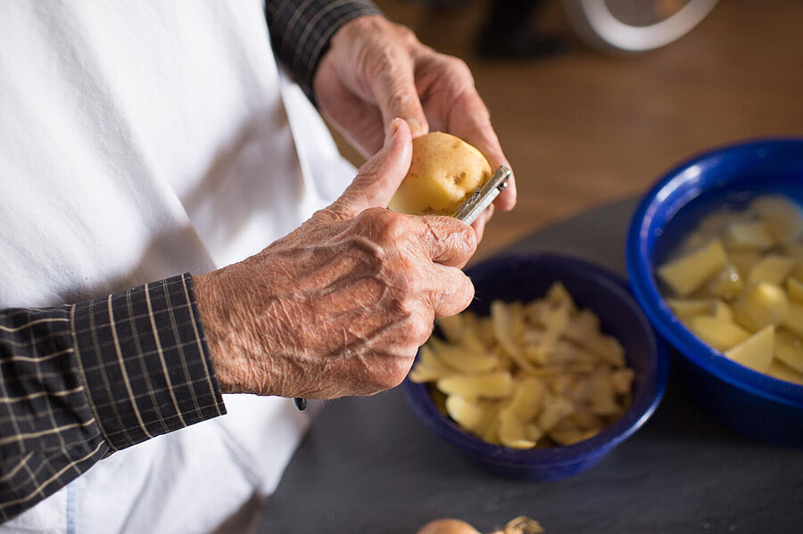 Ein Tisch, zwei blaue Schüsseln. In einer liegen Kartoffelschalen, in der anderen Kartoffelstücke im Wasser. Zwei betagte Hände schälen gerade Kartoffeln.