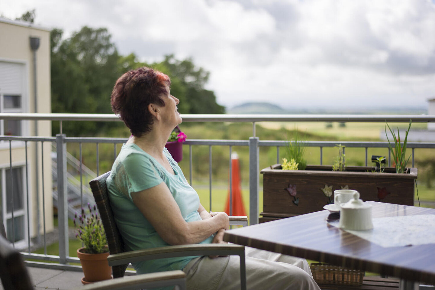 Auf einem Balkon an einem kleinen Tisch eine ältere Dame in hellgrauer Hose und türkisfarbenem Shirt. Sie trinkt Kaffee und schaut  mit erhobenem Kopf in den mit Wolken verhangenen Himmel.
