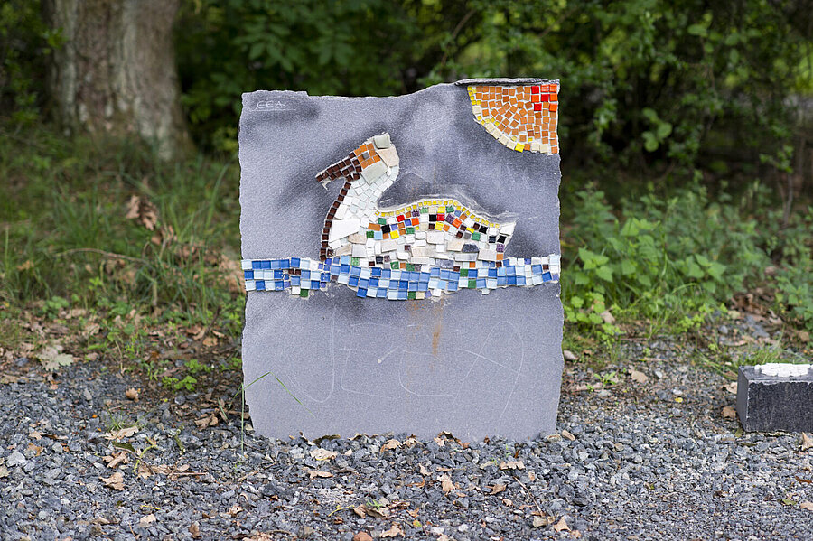 Am Rande einer "wilden" Grundstücksecke mit Schotterweg steht eine glatte, graue, senkrechte Steinplatte. Ein "Künstler" hat mit bunten Glassteinen  eine "Ente auf einem See"gezaubert, die von rechts oben von einer Sonne aus dem gleichen Material beschienen wird.