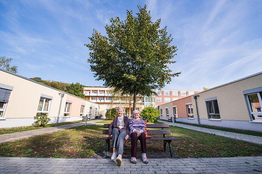 Zwei Seniorinnen sitzen im Innenhof auf einer Bank. Hinter ihnen ein großer, grüner Baum.